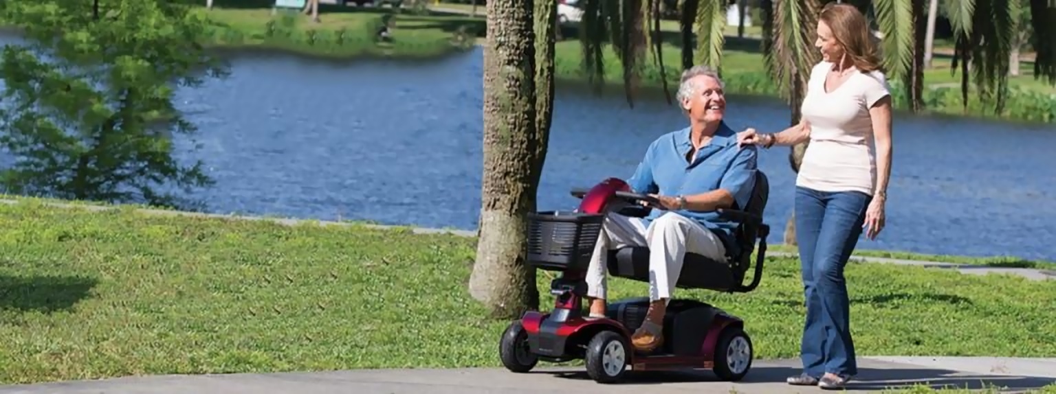 A man on a red mobility scooter beside a companion in a lakeside park